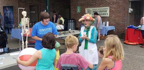 A woman in a floral hat interacts with children near a skeleton display at a community event.