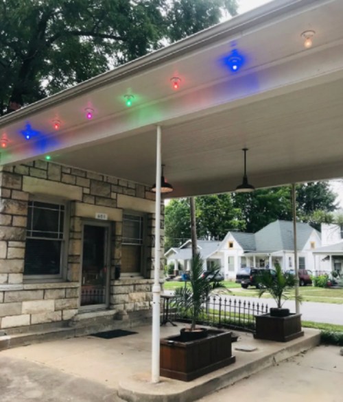 A porch with colorful lights strung along the ceiling, featuring potted plants and a view of nearby houses.