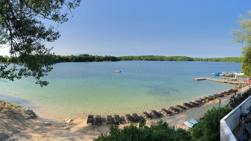 Panoramic view of a calm lake with sandy shore, lounge chairs, and boats docked in the distance under a clear blue sky.