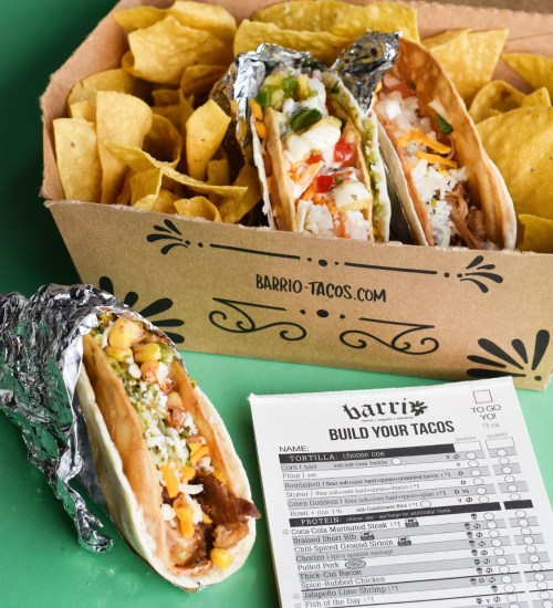 A box of tortilla chips with three tacos, one partially unwrapped, and a taco order form beside them.
