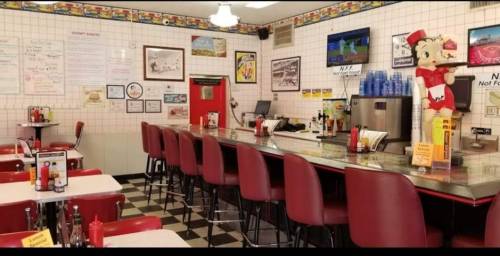 A retro diner interior with red stools, a counter, and vintage decor, featuring a condiment station and a TV.