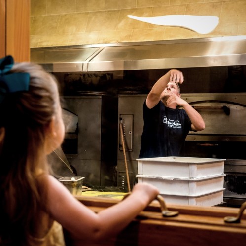 A child watches a chef toss pizza dough in a restaurant kitchen, with steam rising from pots in the background.