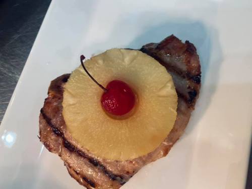 Grilled pork chop topped with a slice of pineapple and a maraschino cherry on a white plate.