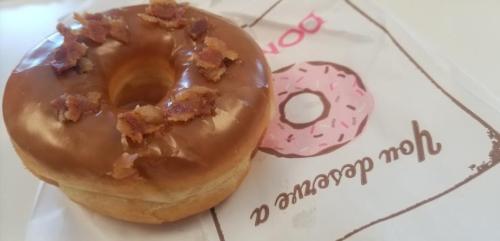 A glazed donut topped with crispy bacon bits, resting on a pink donut-themed paper.