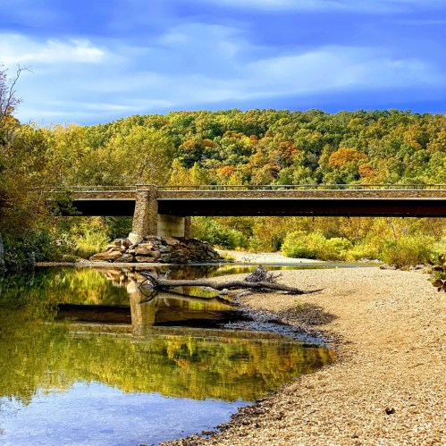 A serene river scene with a bridge, surrounded by colorful autumn trees and a clear blue sky.