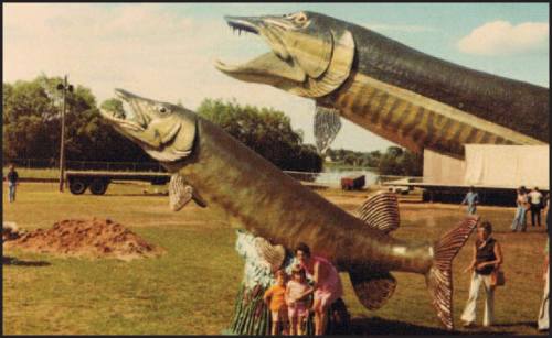 Two large, colorful fish sculptures with open mouths, surrounded by people in a grassy area near a body of water.
