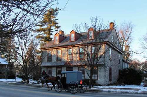 A horse-drawn carriage passes by a historic stone house on a snowy street, with trees in the background.