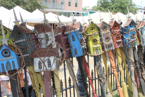 Colorful birdhouses with unique designs, mounted on poles, lined up in a sunny outdoor market setting.