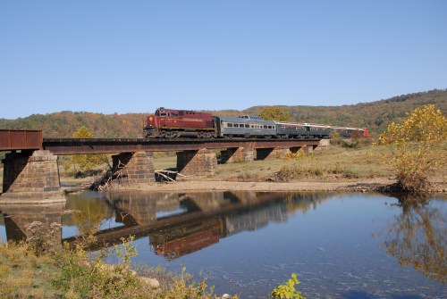 A train crosses a bridge over a calm river, surrounded by autumn-colored trees and clear blue skies.