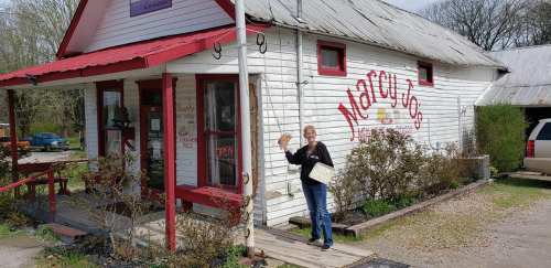 A woman stands outside a quaint, white building with red accents, holding a pie and waving. The sign reads "Mary's."