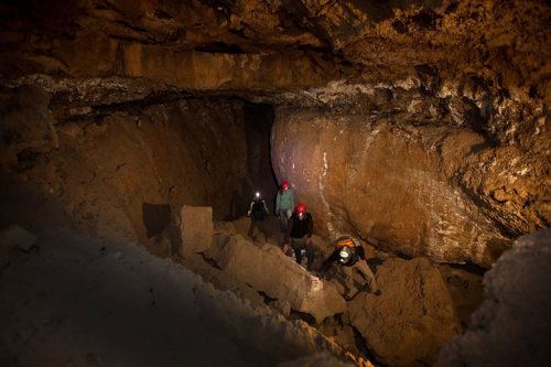 Explorers with headlamps navigate a dark cave, climbing over rocky terrain and illuminated by soft light.