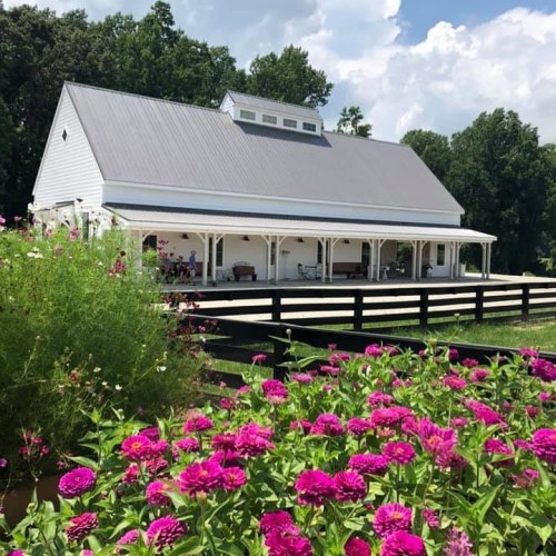 A white barn-style house with a porch, surrounded by vibrant pink flowers and green trees under a blue sky.