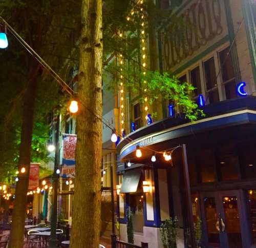 A cozy street scene at night featuring a restaurant with string lights and trees, creating a warm atmosphere.