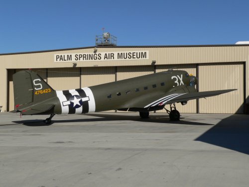 A military aircraft in olive green parked outside the Palm Springs Air Museum, with a clear blue sky in the background.