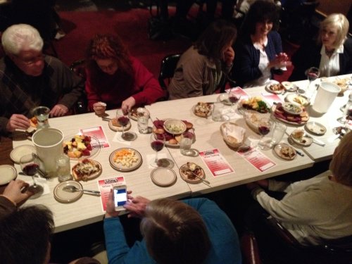 A group of people dining at a long table with plates of food, drinks, and conversation in a cozy setting.