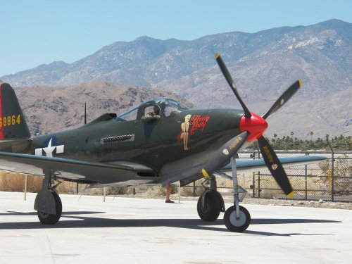 A vintage military aircraft with a pin-up nose art, parked on a tarmac with mountains in the background.