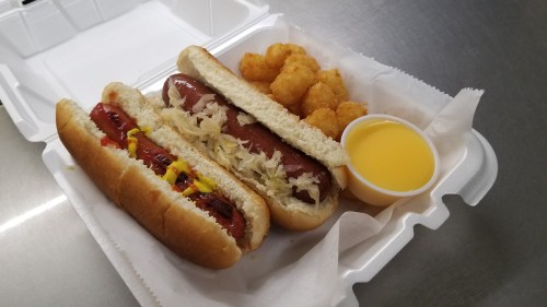 A takeout container with two hot dogs topped with mustard and sauerkraut, tater tots, and a small cup of yellow sauce.