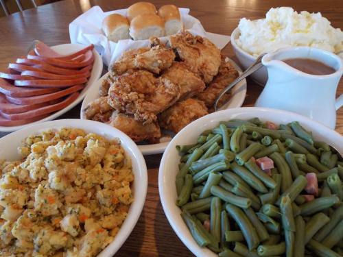 A spread of fried chicken, mashed potatoes, green beans, stuffing, ham slices, and dinner rolls on a wooden table.