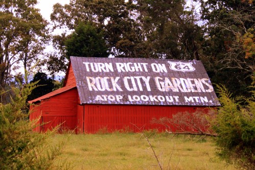 A red barn with a weathered sign reading "Turn Right on Rock City Gardens" atop Lookout Mountain. Surrounded by trees.