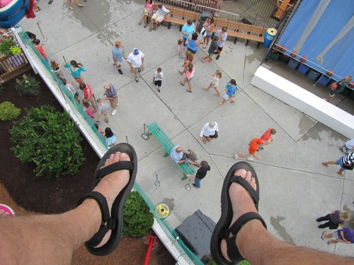 Aerial view of feet in sandals hanging over a crowded amusement park area with people walking below.