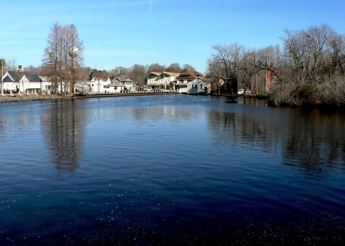 A calm river reflects trees and houses under a clear blue sky, creating a serene landscape.