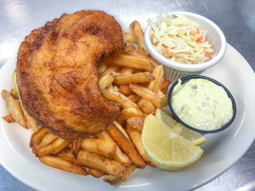 A plate of crispy fried fish with French fries, coleslaw, a lemon wedge, and a side of tartar sauce.