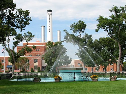 A park with a fountain and trees, featuring a building with "Hastings" and smokestacks in the background.