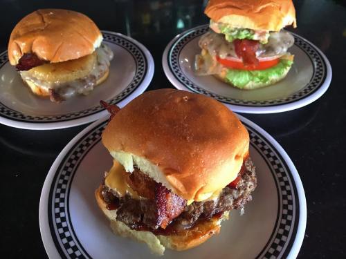 Three gourmet burgers on plates: one with bacon and cheese, one with pineapple, and one with lettuce and tomato.