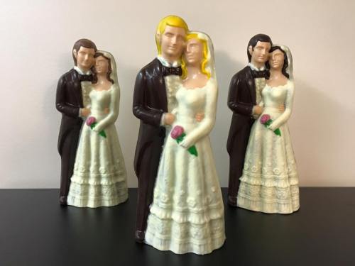 Three ceramic wedding figurines, two in black tuxedos and one in a white dress, holding flowers and embracing.
