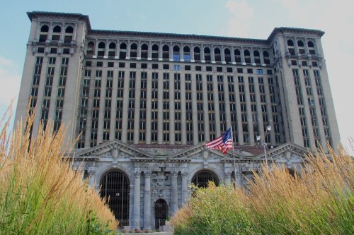 Abandoned historic building with tall windows, overgrown grass, and an American flag in front.