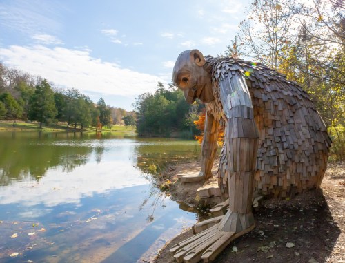 A large wooden sculpture of a giant figure sits by a serene lake, surrounded by trees and a clear blue sky.