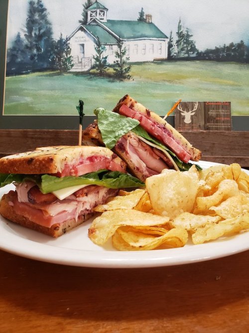 A plate with a stacked sandwich filled with meats and greens, accompanied by a side of potato chips.
