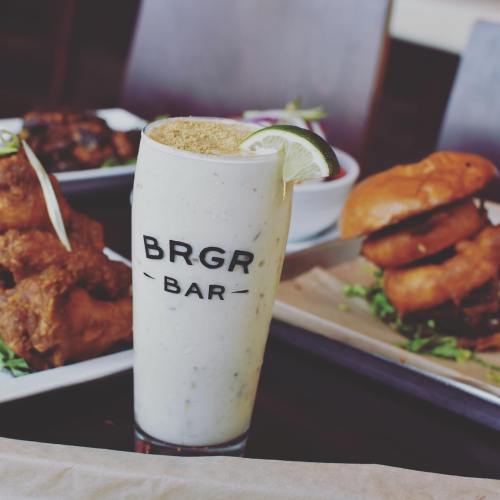 A tall glass of creamy drink with a lime wedge, in front of fried chicken and burgers on a table.