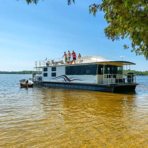 A houseboat on a calm lake with people enjoying the sun on the deck under a clear blue sky.