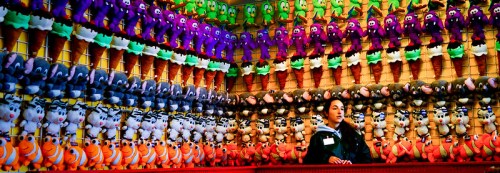 A vendor stands in front of a colorful display of stuffed animals and ice cream cone toys at a fair or carnival.