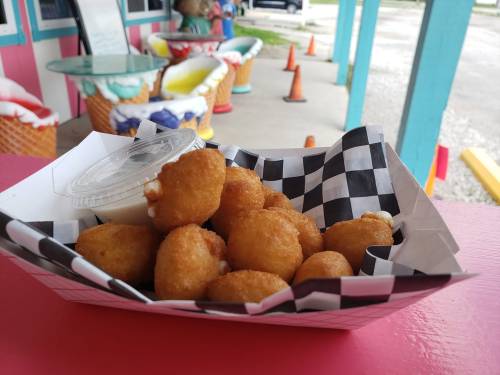 A basket of golden, crispy corn fritters with a small cup of dipping sauce, set against a colorful outdoor backdrop.