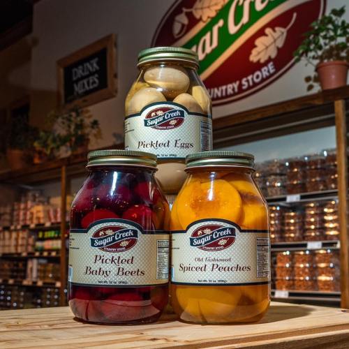 Three jars of pickled foods: pickled eggs, pickled baby beets, and spiced peaches, displayed in a country store.
