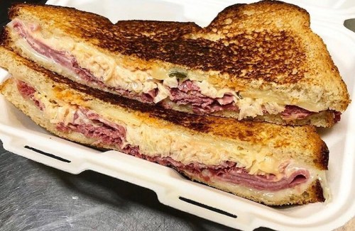 A toasted sandwich cut in half, filled with layers of ham, cheese, and sauerkraut, served in a takeout container.