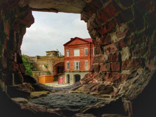 View through a stone wall opening, showcasing a red brick building and a cloudy sky in the background.