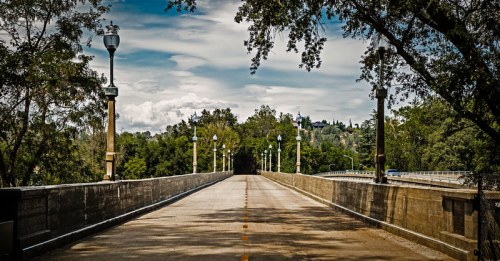 A wide, empty bridge lined with trees and street lamps, leading into a lush green landscape under a cloudy sky.
