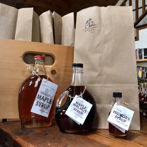 Three bottles of syrup (maple, maple walnut, hickory) displayed next to a paper bag in a rustic setting.