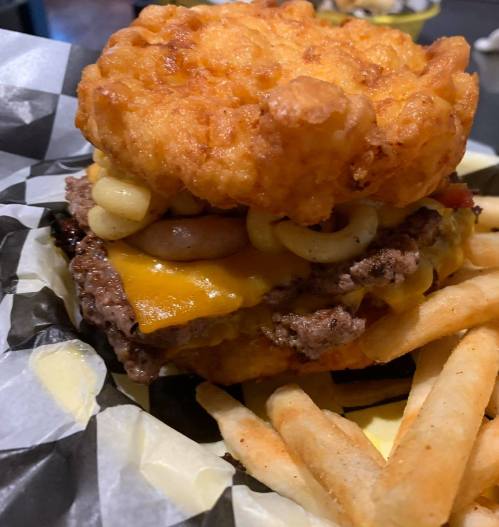 A close-up of a hearty burger with a fried mac and cheese bun, cheese, and fries on the side.