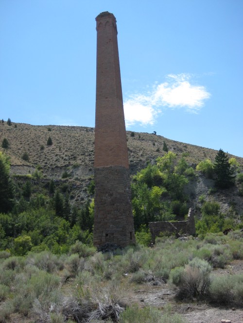 A tall, brick chimney stands alone in a rugged landscape, surrounded by shrubs and hills under a clear blue sky.