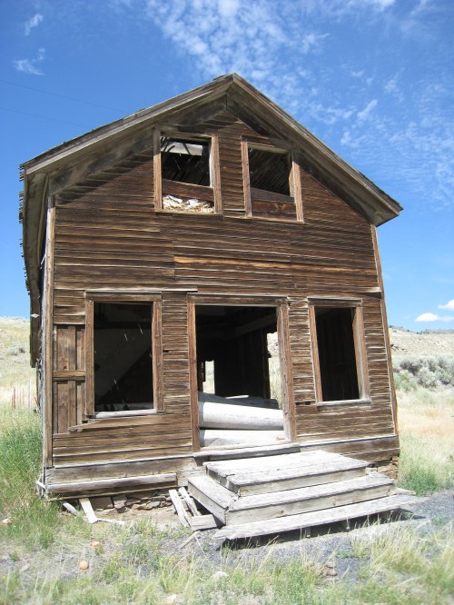 Abandoned wooden house with broken windows and a slanted roof, set against a blue sky and grassy landscape.