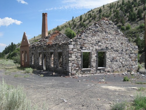 Ruins of a stone building with a chimney, surrounded by greenery and mountains under a clear blue sky.