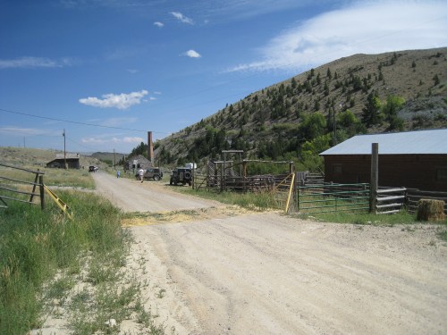 A dirt road leads to a rustic farm with wooden buildings and a mountainous backdrop under a blue sky.