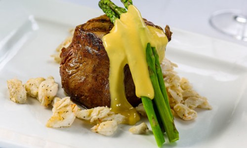 A gourmet dish featuring a tender steak topped with hollandaise sauce and asparagus, served on a bed of crumbled bread.