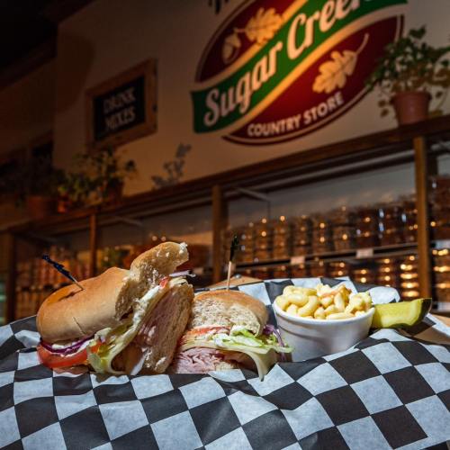A delicious sandwich with lettuce, tomato, and ham, served with macaroni and cheese and a pickle on a checkered plate.