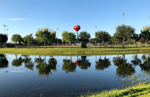 A serene park scene with a water tower, trees, and reflections in a calm pond under a clear blue sky.