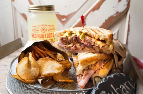A Cuban sandwich with layers of meat and cheese, served with crispy potato chips and a jar of lemonade.